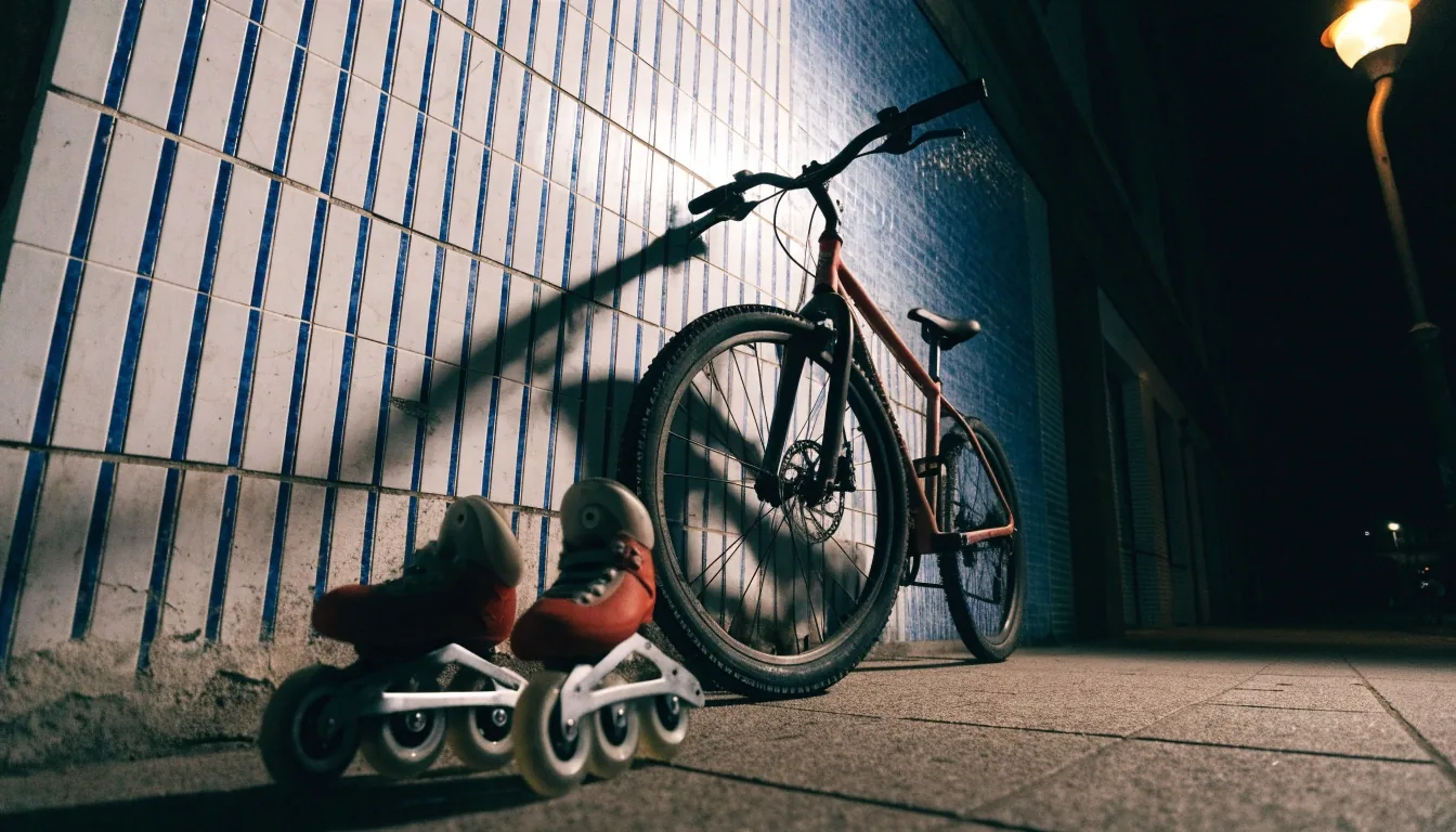 Bicicleta usada apoyada en una pared de azulejos con patines en línea junto a ella, iluminada por luz de farola
